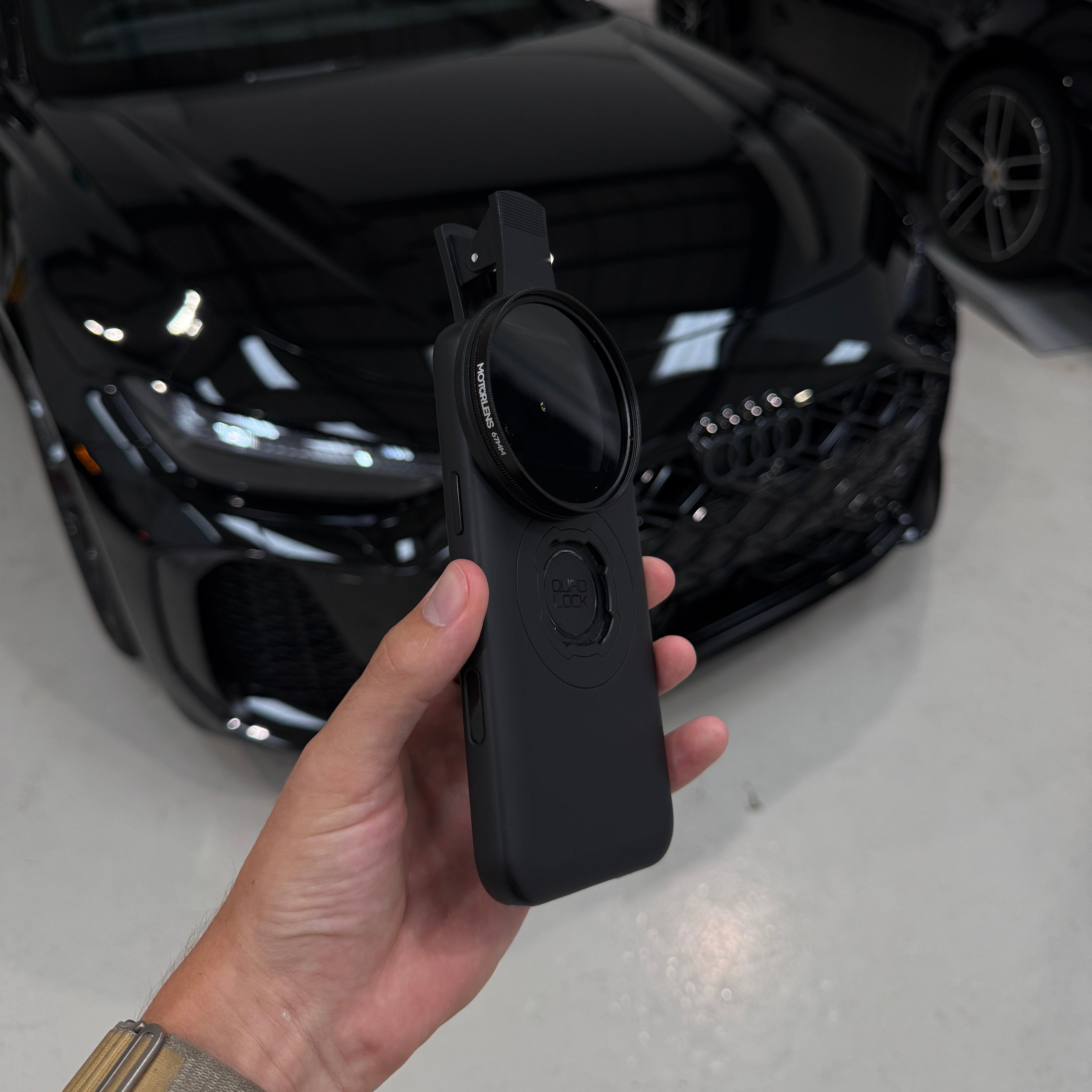 A hand holding a black circular lens filter with a rectangular mounting case, with a glossy black car and a garage floor in the background.