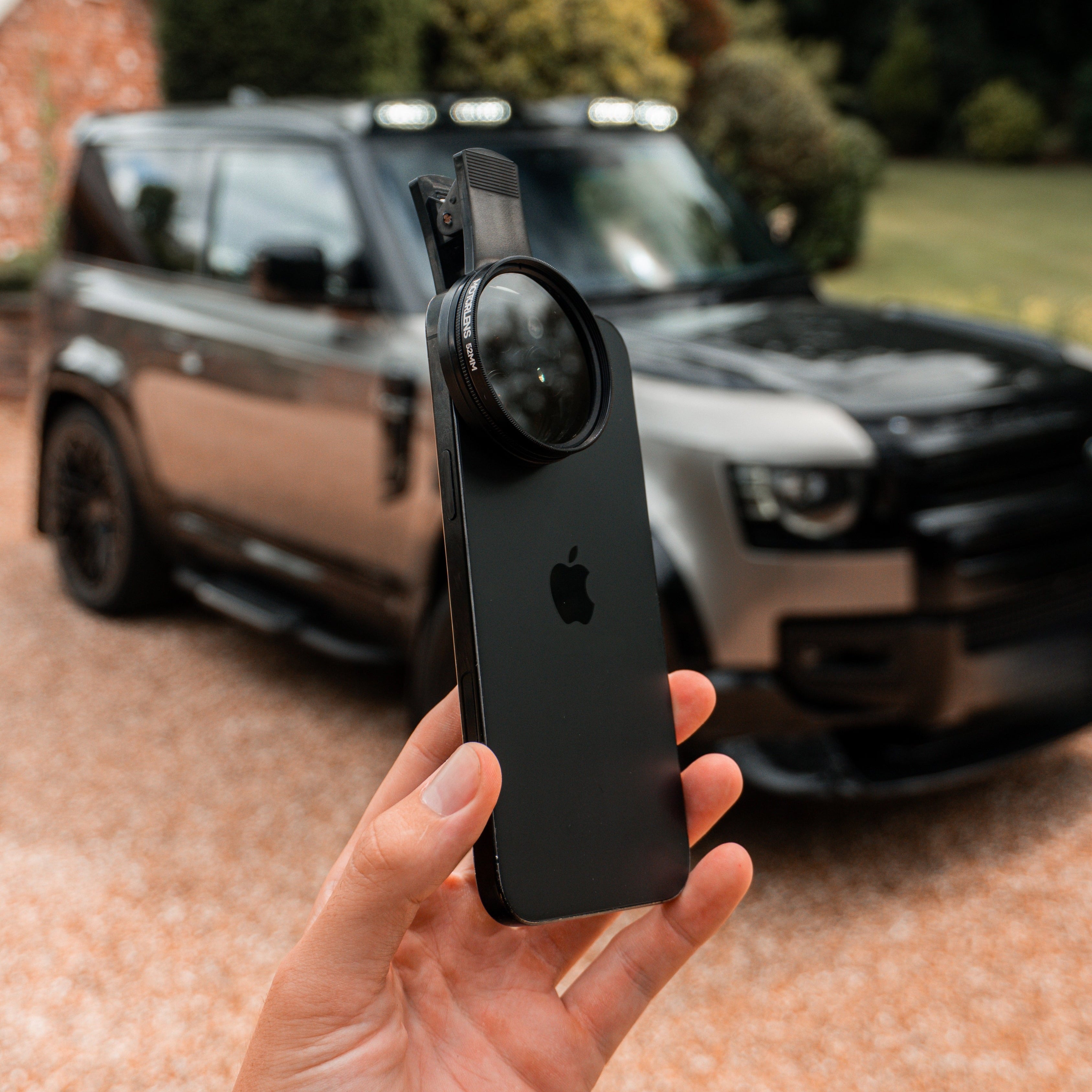Person holding a smartphone with a camera lens attachment in front of a car
